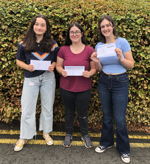 Three students stood infront of a bush holding their exam results on paper.