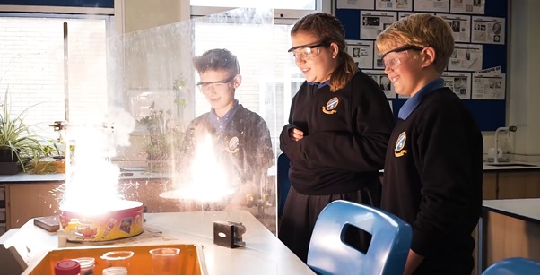 Three students behind a safety screen observing a Science experiment burning very brightly